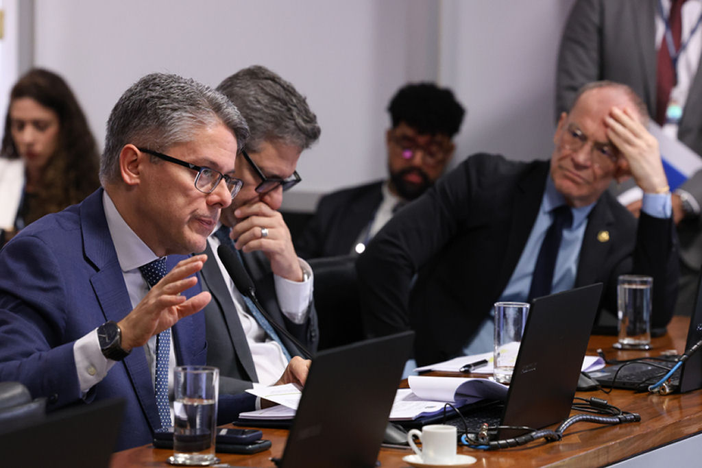 Homem de terno e óculos falando ao microfone durante reunião de comissão governamental, cercado por notebooks e outros participantes.