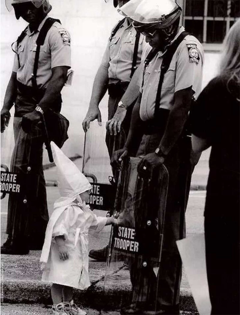 Na fotografia vê-se a uma menina vestida com o uniforme tradicional do KKK tocando seu reflexo no escudo de um polícial afro-americano em 1992.